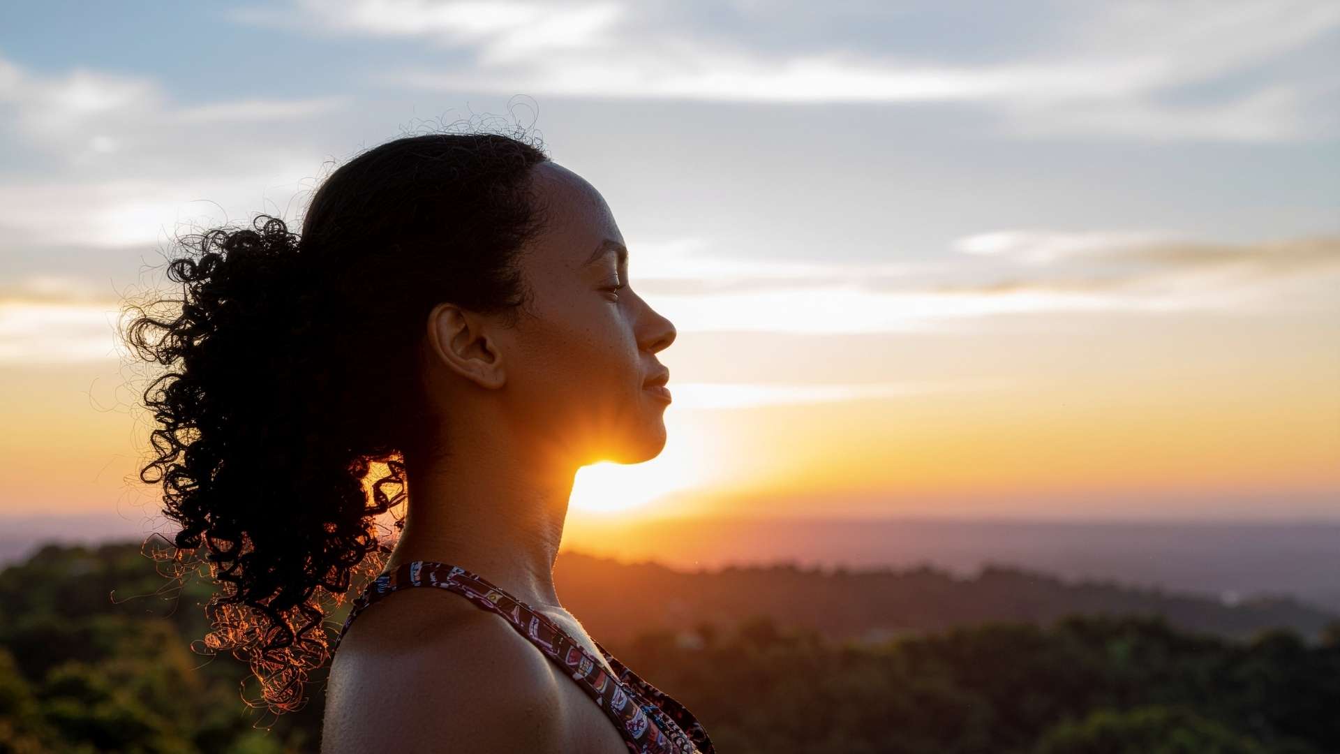 A contemplative-looking woman standing on an overlook as the sun sets in the background.