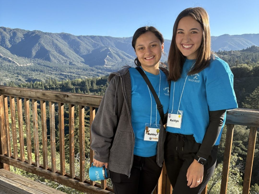 Ashley Gonzalez and Kaitlyn Saunders, Loma Linda University Child Life Specialist MS students, smile on a wooden deck overlooking a mountain landscape during Camp Good Grief.