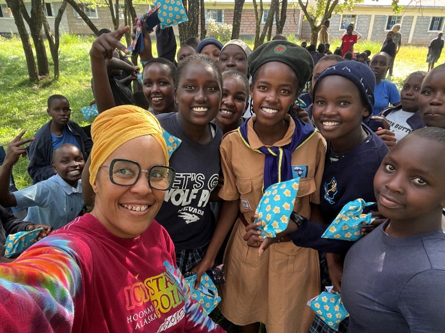 Akiliah Perez smiles for a selfie with a group of Kenyan schoolgirls holding small blue gift pouches, with more children gathered outdoors behind them.