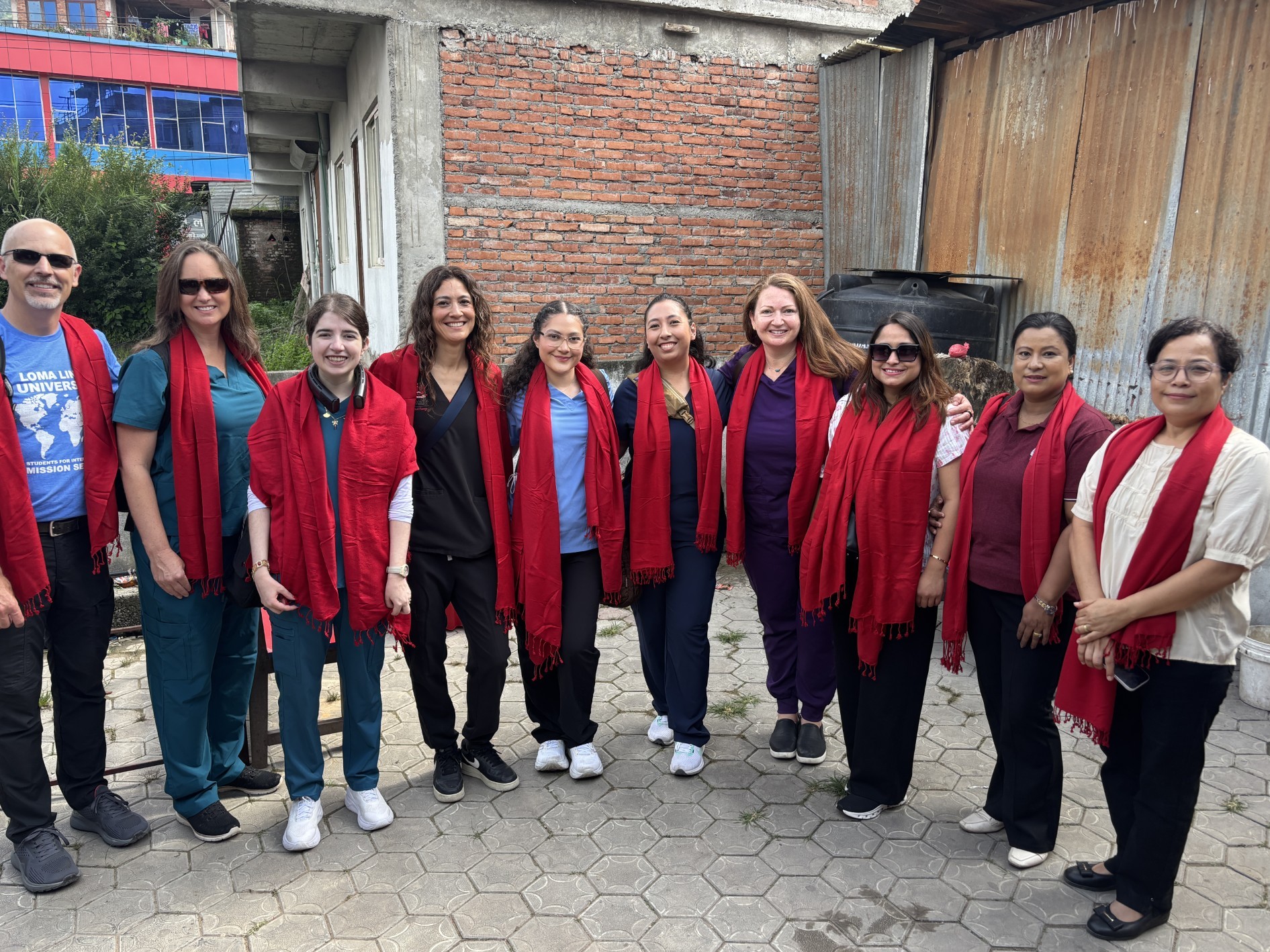 Ten people in scrubs and casual clothing stand in a row in a paved courtyard, each wearing a red scarf, in front of a brick wall and a corrugated metal fence. One man wears a Loma Linda University Mission Service t-shirt.