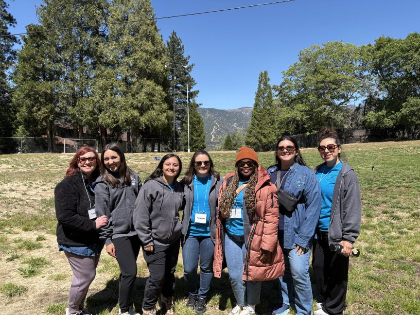 A group of seven Camp Good Grief counselors smile together on an open field surrounded by tall pine trees and mountain views.