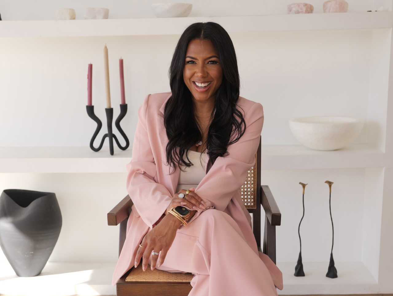 Dr. Allycin Powell-Hicks smiling in a blush pink suit, seated in a cane-back armchair in a styled, modern interior.