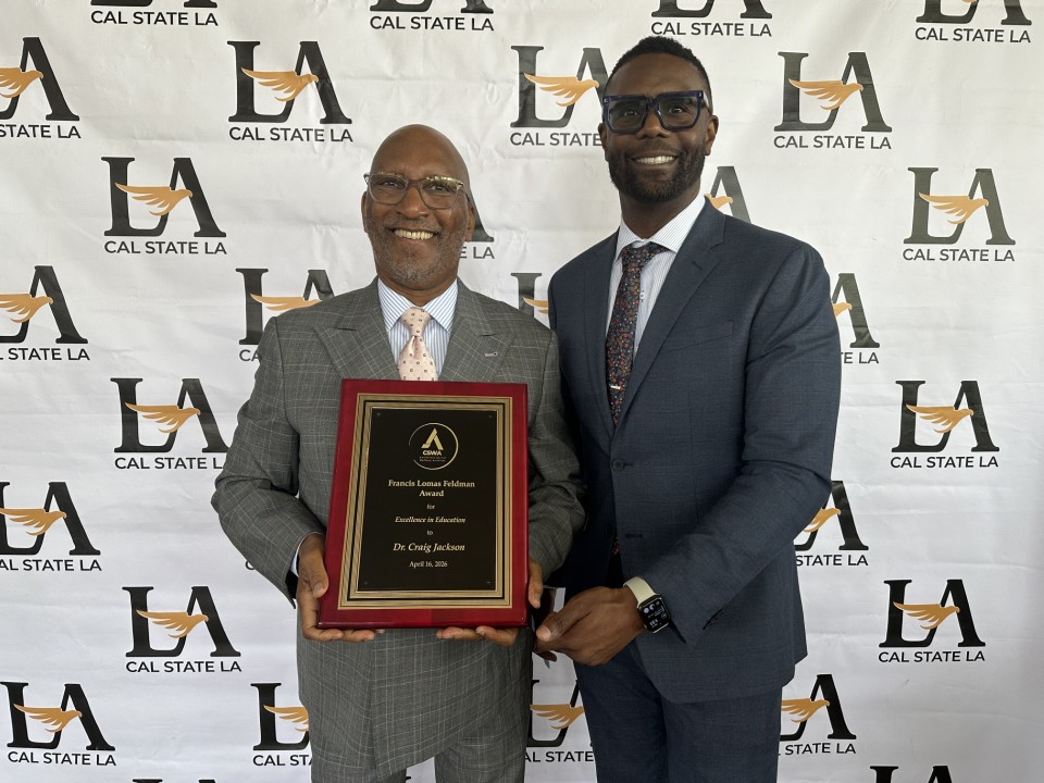 Dr. Craig Jackson and Dr. Maurice Hudson stand together in front of a logo step and repeat background, smiling at the camera. Dr. Jackson, on the left, is holding an award. Both are wearing suits and ties.