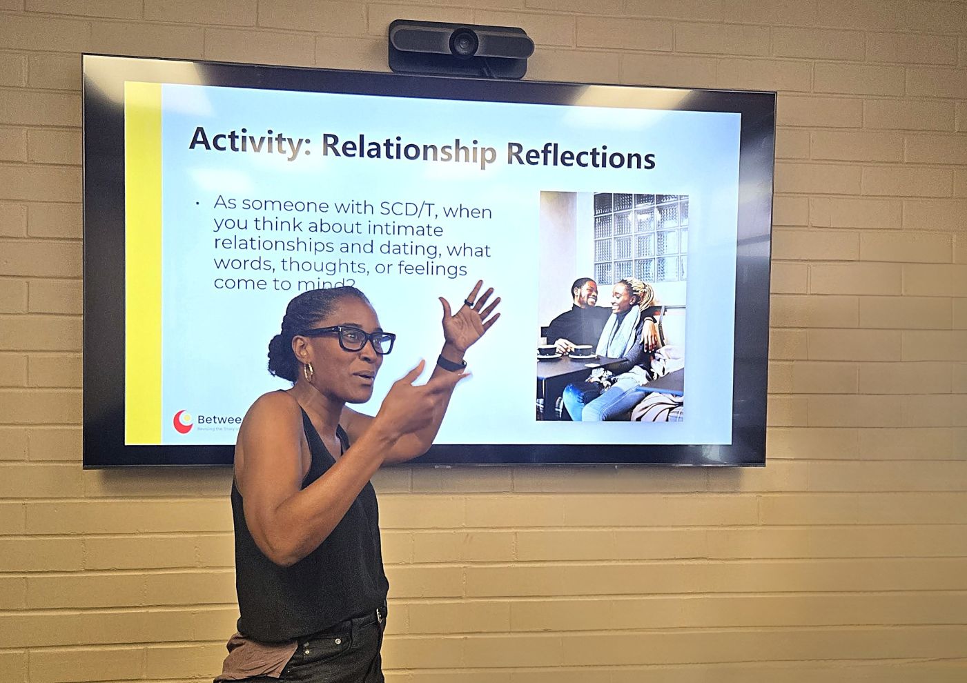 Dr. Monique Willis presenting a slide titled 'Activity: Relationship Reflections' during a talk on relationships and dating for people with sickle cell disease and trait, with a photo of a couple smiling together displayed on screen.