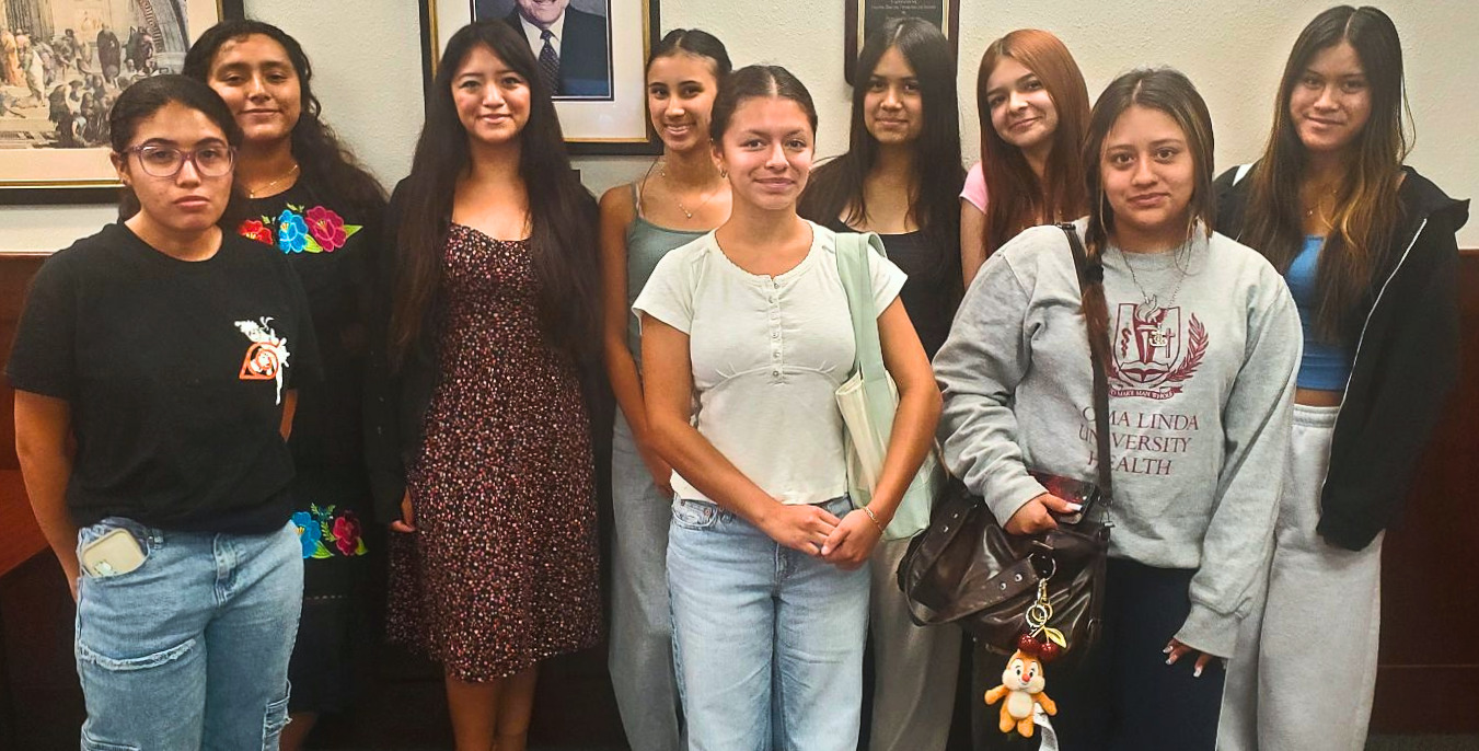 Group of nine female students smiling together in a wood-paneled office, standing in front of framed portraits on the wall.