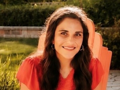 Dr. Lorraine Benuto, a woman with long dark wavy hair, smiling while seated in an orange Adirondack chair outdoors, wearing a red top and hoop earrings, with greenery in the background.