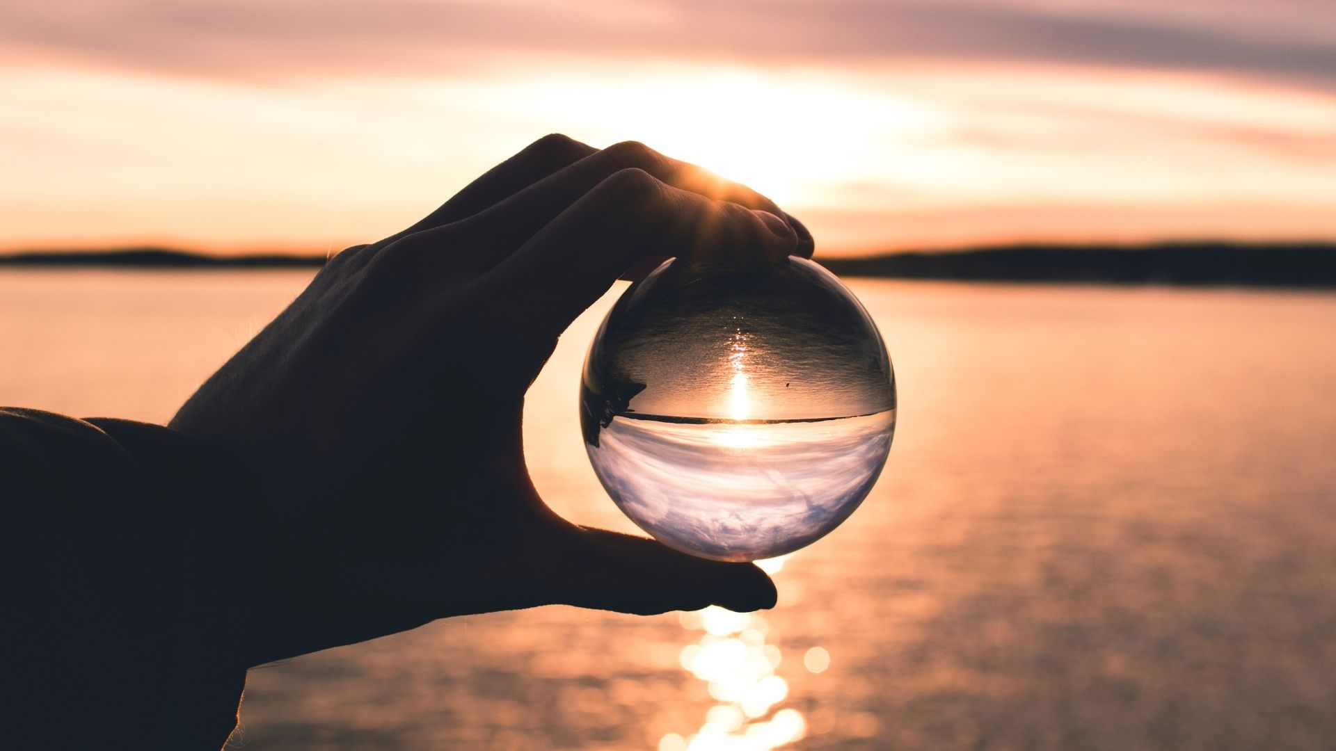 A hand cradles a glass orb reflecting a peaceful sunset over calm water, symbolizing clarity, reflection, and inner calm.