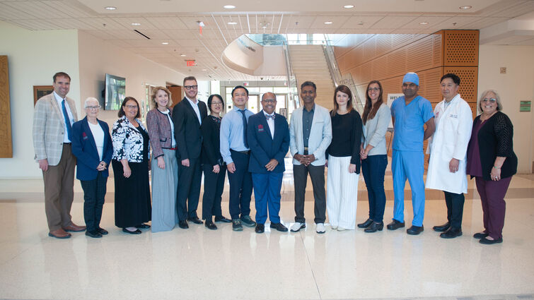 A group of 14 researchers and faculty in business attire, scrubs, and a white coat pose together in a bright hospital lobby with a spiral staircase in the background.