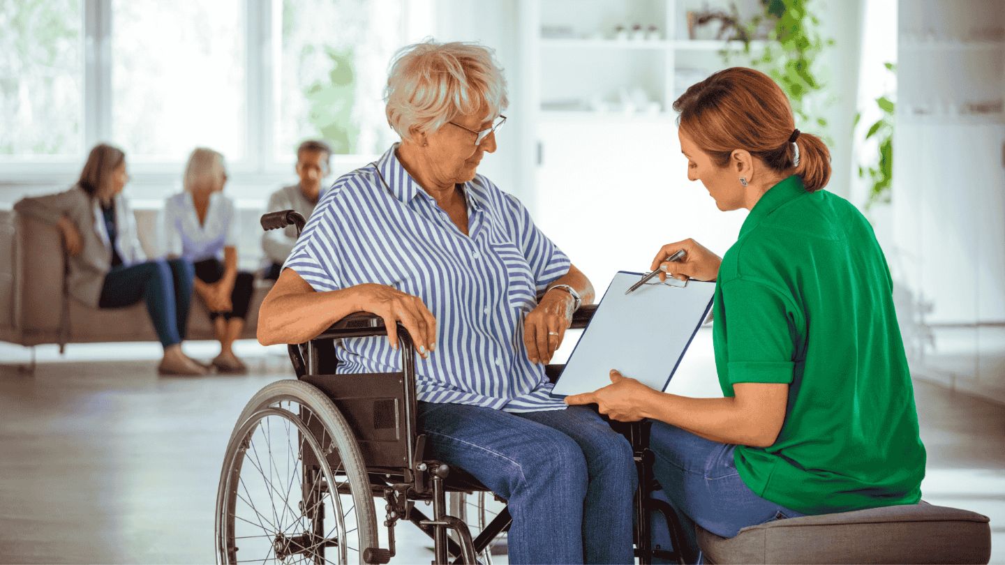 An older woman in a wheelchair speaks with a younger woman holding a clipboard