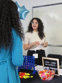 Two Loma Linda University Human Resources representatives speak with a student at their booth during the 2026 SBH Job Fair. The table displays a Connect Four game, candy, and informational materials including a 2025 Facts and Figures sheet.