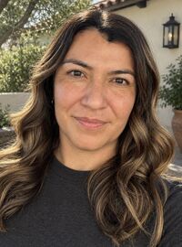 Close-up headshot of Dr. Danielle Lascano with long wavy brown hair smiling softly outdoors, with greenery and a stone building in the background.