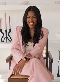 Dr. Allycin Powell-Hicks smiling in a blush pink suit, seated in a cane-back armchair in a styled, modern interior.