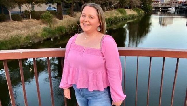 Student Alice Donovan smiles at the camera in front of a railing at dusk, wearing a pink shirt and jeans. Behind her is a body of water with docks, buildings, and trees visible.