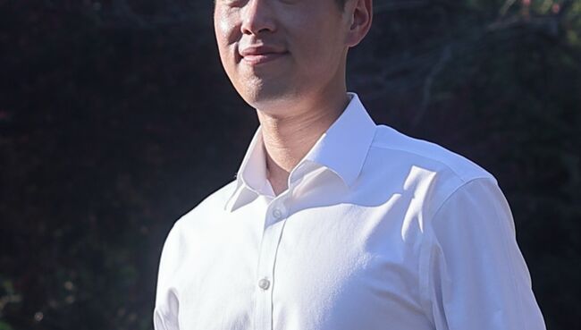 Student Jacob Lui looks off-camera and smiles. He is standing on a bridge and wearing business casual attire. Trees are visible in the background.