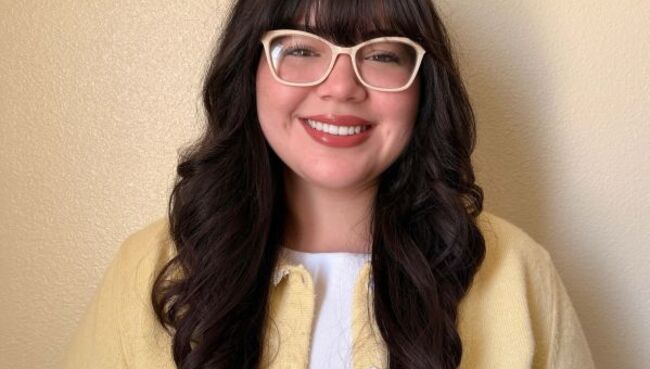 Student Karly Franco smiles at the camera, wearing glasses, a white shirt, and a pale yellow cardigan. The background is a pale yellow wall.