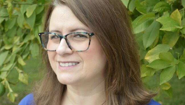 Student Olivia Seheult smiles at the camera, wearing glasses and a blue shirt. In the background are green leaves.