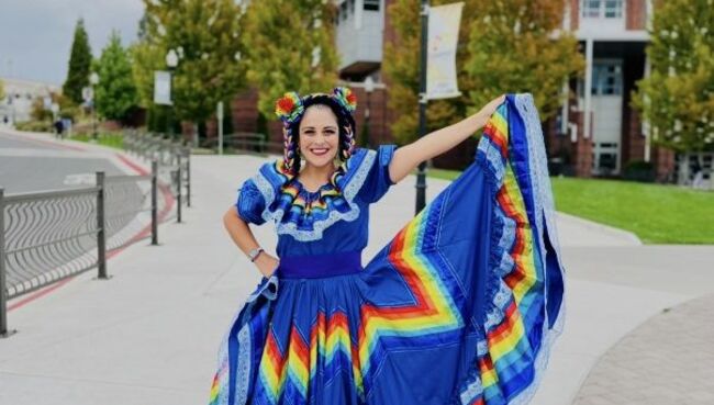 Dr. Lorraine Benuto wears a vibrant blue traditional Mexican folklórico dress with a rainbow-striped skirt and matching hair ribbons, smiling and holding out the skirt on a campus walkway with brick buildings and trees in the background.