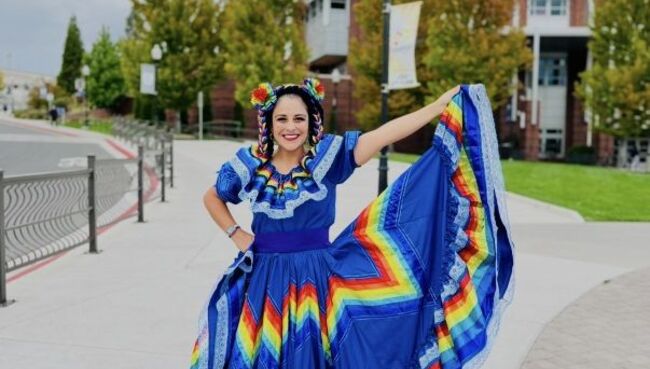 Dr. Lorraine Benuto wears a vibrant blue traditional Mexican folklórico dress with a rainbow-striped skirt and matching hair ribbons, smiling and holding out the skirt on a campus walkway with brick buildings and trees in the background.