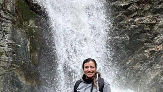 Dr. Danielle Lascano smiling at the camera while standing in front of a waterfall, wearing a gray hoodie and backpack, surrounded by rocky terrain.