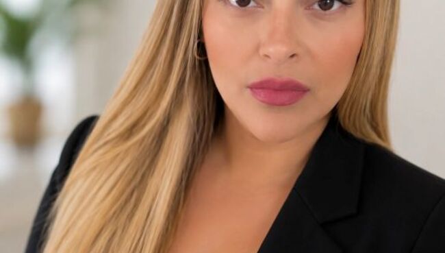 Professional headshot of online MSW student Isaura Alvarez Nepomuceno, a woman with long, straight, honey-blonde hair, wearing a black blazer over a white top and a rose-colored lipstick, in a softly lit, bright indoor setting.
