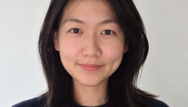 Portrait of Counseling MS student Julie Ma with shoulder-length dark hair parted in the center, wearing a navy sweater and a soft, closed-mouth smile, photographed in front of a plain white wall.