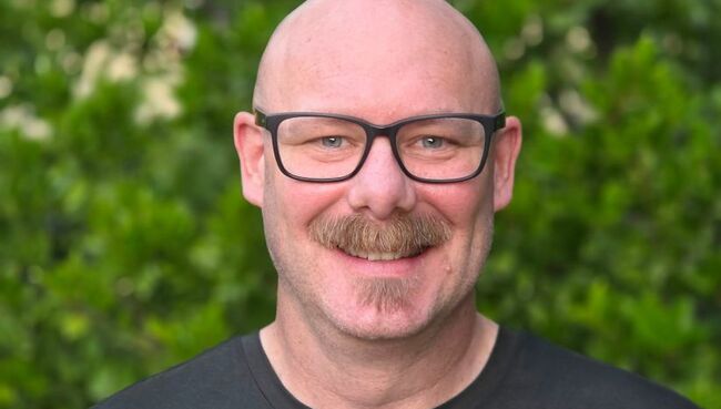 Pat Keplinger, a student in the MFT MS on-campus program, smiling warmly at the camera. He wears dark-framed glasses and a dark gray t-shirt and is pictured outdoors in front of lush green foliage with small white flowers.