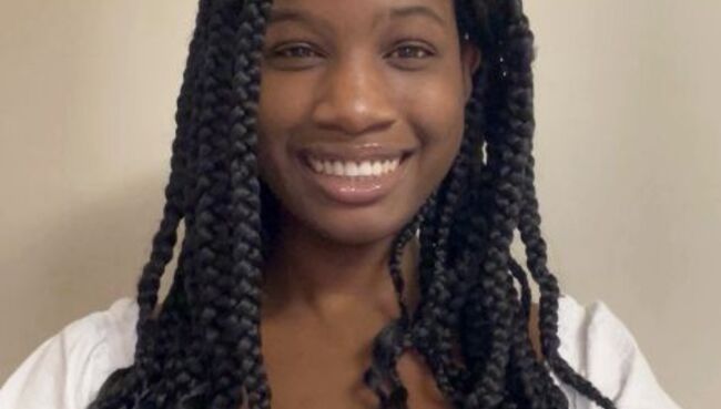 Serene Oler, an MSW advanced standing student, smiling warmly at the camera. She has long black braids and wears a white top with short puffed sleeves, pictured against a neutral beige wall.