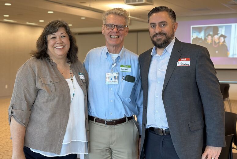 Prof. Michelle Minyard-Widmann, Prof. Randall Walker, and Dr. Brian Distelberg standing together and smiling with a projected PowerPoint presentation visible in the background