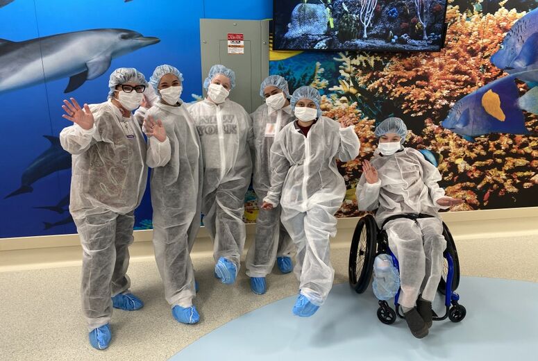 Child Life students in surgical masks, shoe covers, and other personal protective equipment posed as a group in front of an ocean-themed wall at the Loma Linda University Health Surgical Hospital