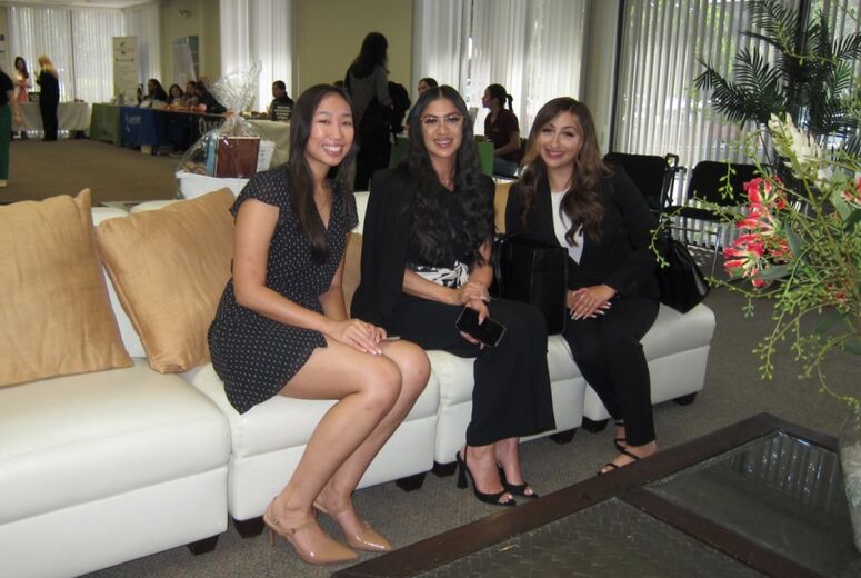 Three professionally dressed students smile for a photo together on a white couch in the job fair venue, with employer booths and other attendees visible in the background.