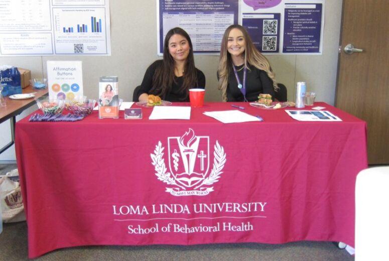 Two staff sit behind a table draped with a maroon Loma Linda University School of Behavioral Health tablecloth, with affirmation buttons and other promotional materials displayed at their booth.