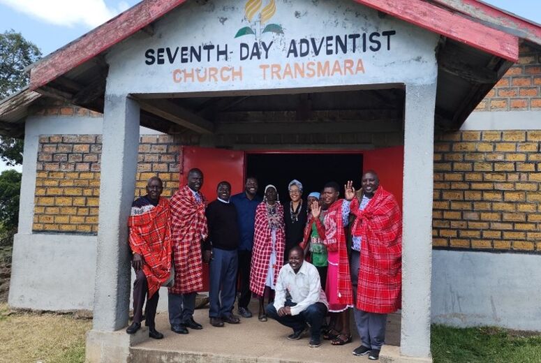 Akiliah Perez poses with a group of Maasai men and women, several wearing traditional red plaid shawls, outside the entrance of the Transmara Seventh-day Adventist Church, a brick building with a red roof.