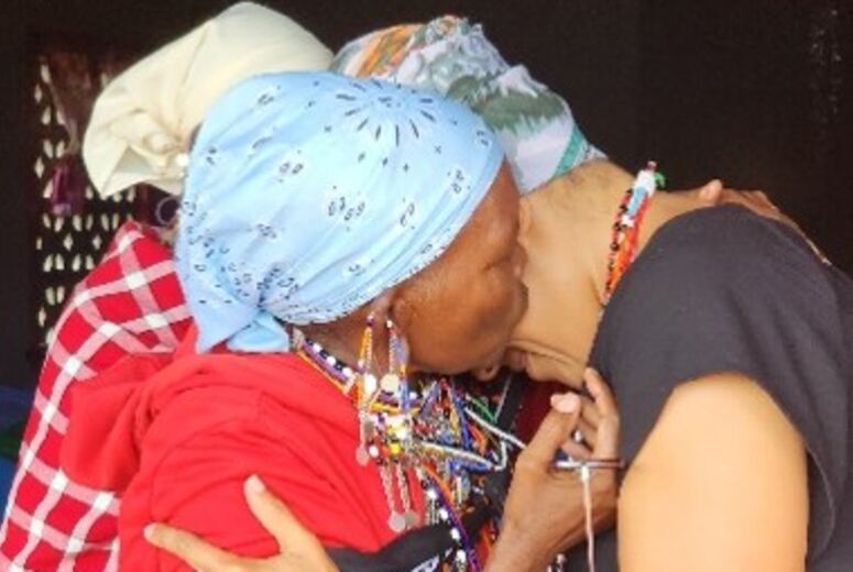 A Maasai woman in a red dress, blue headscarf, and beaded jewelry embraces Akiliah Perez in a warm hug beneath the church's covered entrance.