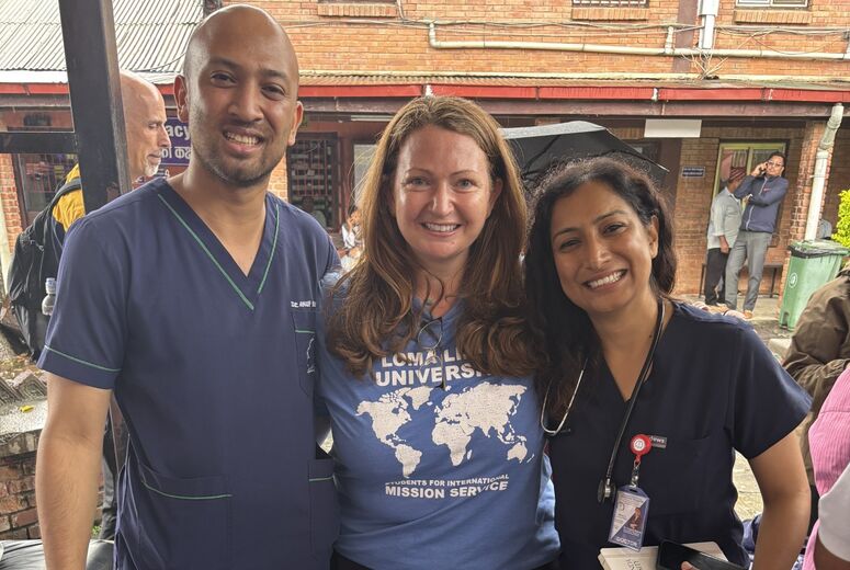A woman in a blue Loma Linda University Students for International Mission Service t-shirt poses between two doctors in navy scrubs, outdoors in front of a brick hospital building.