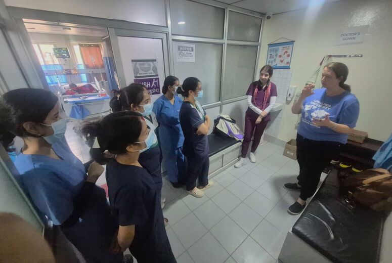 Nurses in scrubs and face masks stand in a hospital corridor listening to a woman in a blue Loma Linda University Mission Service t-shirt, who gestures as she speaks. An intensive care bed is visible through glass doors behind them.