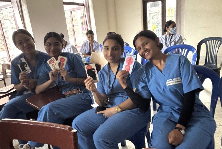 Four smiling students in blue scrubs sit together in a classroom, holding up illustrated cards. One card reads “Leadership.”
