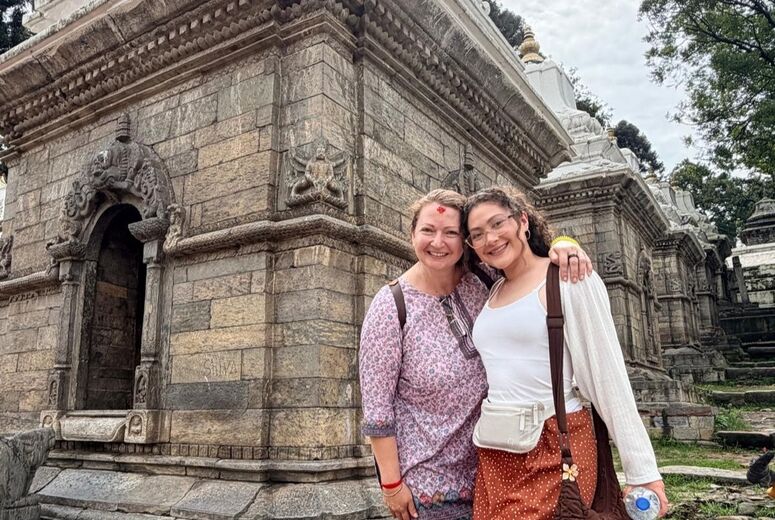 Two women smile and pose arm in arm in front of a carved stone temple. One wears a pink floral tunic with a red tika on her forehead, and the other wears glasses, a white top, and a rust polka dot skirt.