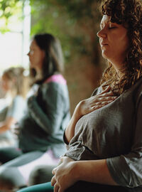 A pregnant woman sits with her eyes closed, one hand on her chest and one hand on her belly, practicing mindfulness in a group setting.