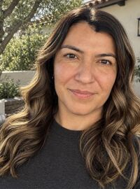 Close-up headshot of Dr. Danielle Lascano with long wavy brown hair smiling softly outdoors, with greenery and a stone building in the background.