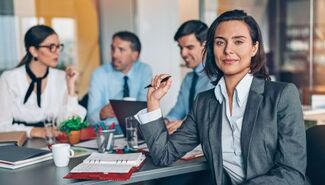 Woman in a gray blazer sitting at a conference table holding a pen, with three colleagues in business attire in a discussion behind her.