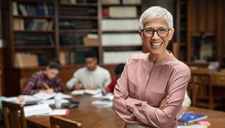 Smiling woman with short gray hair and glasses standing with arms crossed in a library, with students studying at a table behind her.