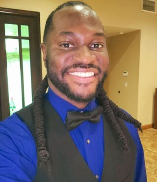 Student Maurice Williams smiles at the camera, wearing a bright blue shirt, black vest, and black bowtie. In the background, some yellow walls and a door are visible.