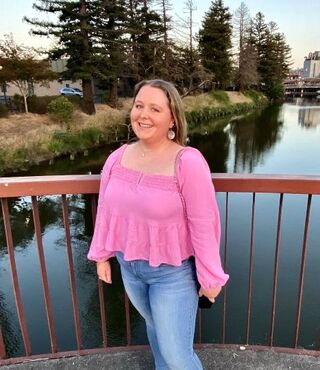 Student Alice Donovan smiles at the camera in front of a railing at dusk, wearing a pink shirt and jeans. Behind her is a body of water with docks, buildings, and trees visible.