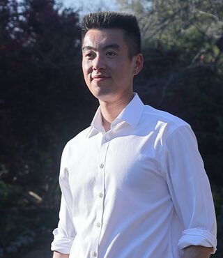 Student Jacob Lui looks off-camera and smiles. He is standing on a bridge and wearing business casual attire. Trees are visible in the background.