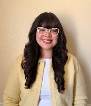 Student Karly Franco smiles at the camera, wearing glasses, a white shirt, and a pale yellow cardigan. The background is a pale yellow wall.