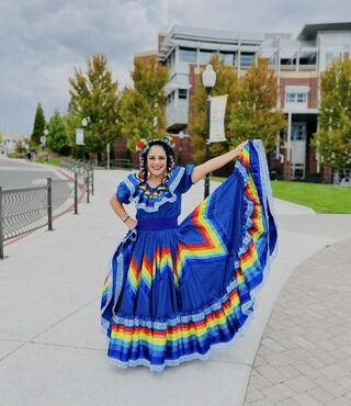 Dr. Lorraine Benuto wears a vibrant blue traditional Mexican folklórico dress with a rainbow-striped skirt and matching hair ribbons, smiling and holding out the skirt on a campus walkway with brick buildings and trees in the background.