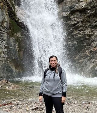 Dr. Danielle Lascano smiling at the camera while standing in front of a waterfall, wearing a gray hoodie and backpack, surrounded by rocky terrain.