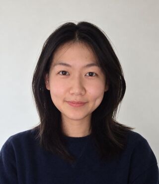 Portrait of Counseling MS student Julie Ma with shoulder-length dark hair parted in the center, wearing a navy sweater and a soft, closed-mouth smile, photographed in front of a plain white wall.