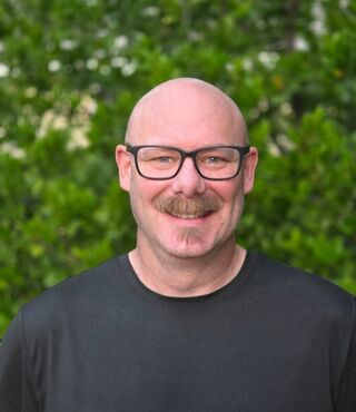 Pat Keplinger, a student in the MFT MS on-campus program, smiling warmly at the camera. He wears dark-framed glasses and a dark gray t-shirt and is pictured outdoors in front of lush green foliage with small white flowers.