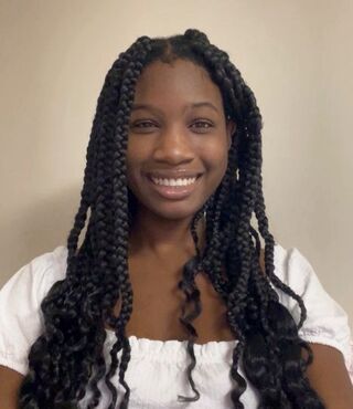 Serene Oler, an MSW advanced standing student, smiling warmly at the camera. She has long black braids and wears a white top with short puffed sleeves, pictured against a neutral beige wall.